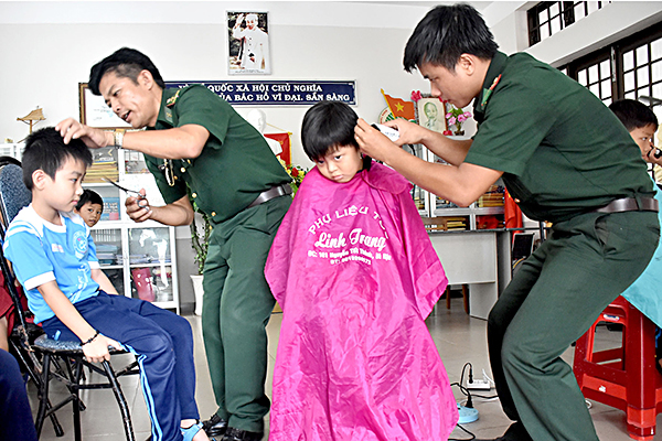 Trung uý Vũ Thanh Tùng (bìa phải), Đội trưởng Đội tham mưu hành chính Đồn Biên phòng Phước Tỉnh cắt tóc miễn phí cho HS Trường TH Lê Hồng Phong (huyện Long Điền) trong chương trình “Tay kéo Biên phòng”.