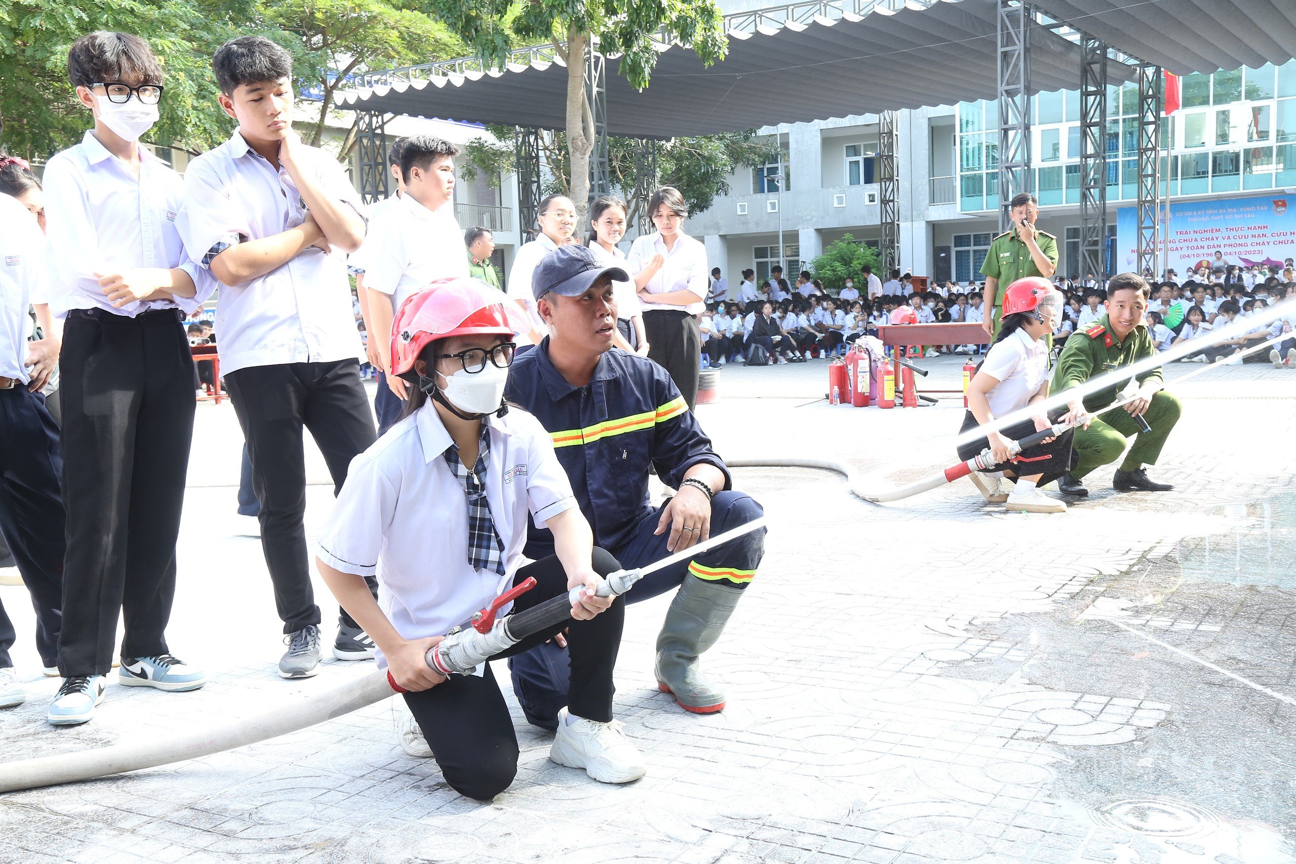 Học sinh cầm lăng vòi chữa cháy, được hướng dẫn cách sử dụng để phun nước tiêu điểm dập tắt đám cháy.
